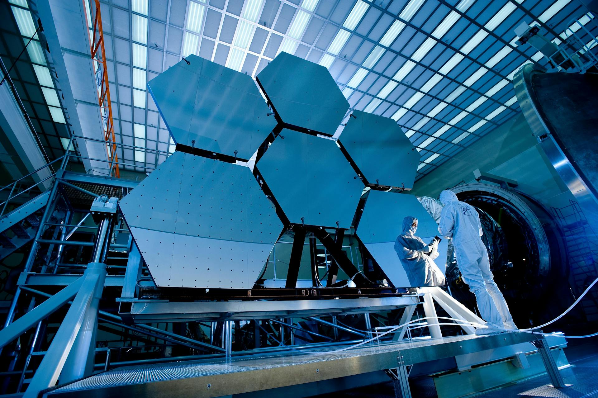 A person in cleanroom suits inspect a large hexagonal mirror assembly in a high-tech laboratory with metal structures and overhead lighting.