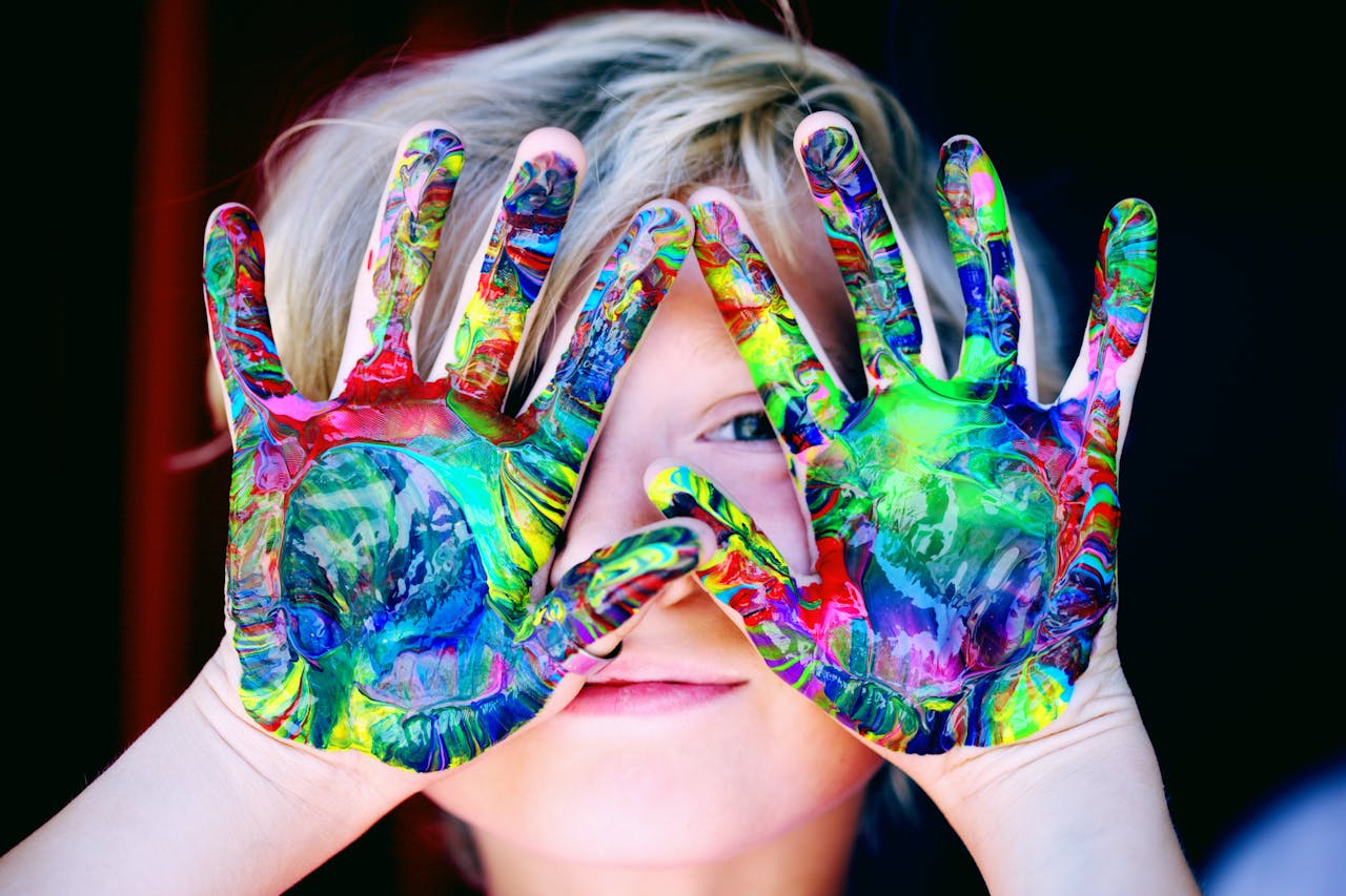 A young child hides their eyes with hands covered in vibrant paint, showcasing a moment of imaginative play